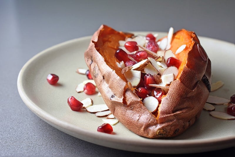 Side view of a baked sweet potato on a plate topped with toasted almond slices and pomegranate arils.