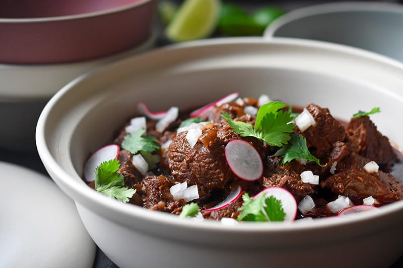 Side view of Instant Pot Cowboy Chili in a white ceramic serving bowl, topped with cilantro, sliced radishes, and diced white onions.