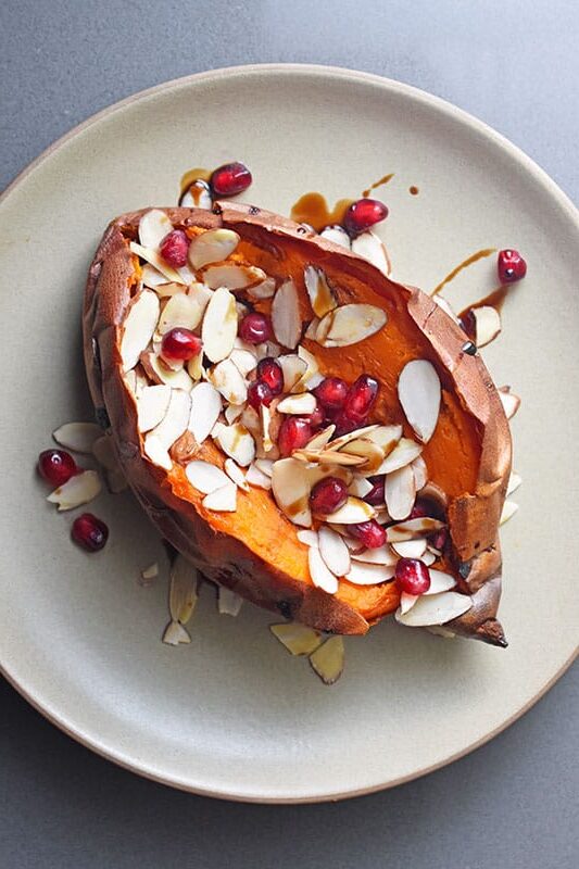 Overhead view of a baked sweet potato on a plate topped with toasted almond slices and pomegranate arils.