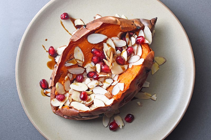 Overhead view of a baked sweet potato on a plate topped with toasted almond slices and pomegranate arils.