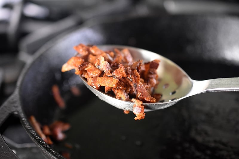 Removing the crispy bacon bits with a slotted spoon from a cast iron skillet