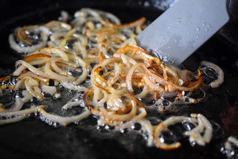 A close up of thinly sliced shallots frying in a cast iron skillet until soft and browned.