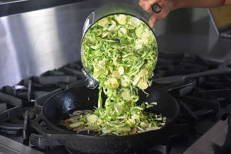 Freshly shaved Brussels sprouts is poured from a food processor container into a large cast iron skillet.