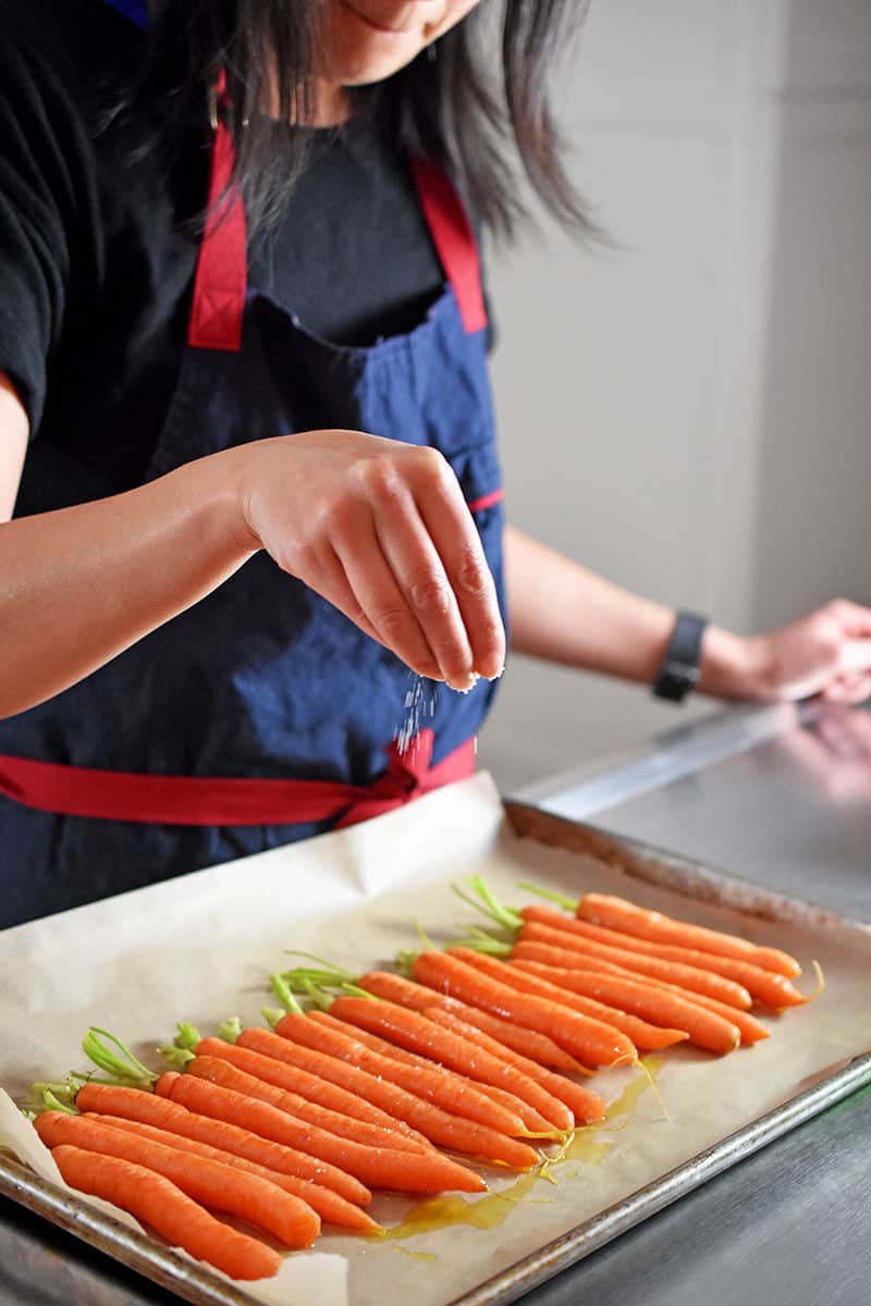 A black haired woman is sprinkling salt on a tray of carrots.