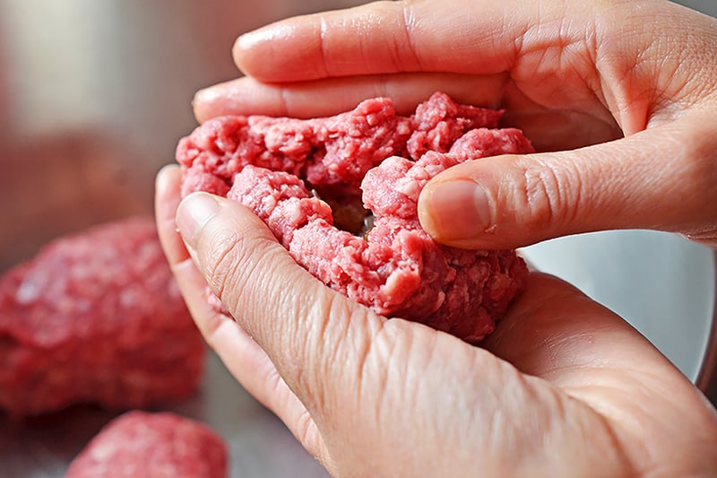 Two hands are sealing up the ground beef around the cooked shallots to form a ball.