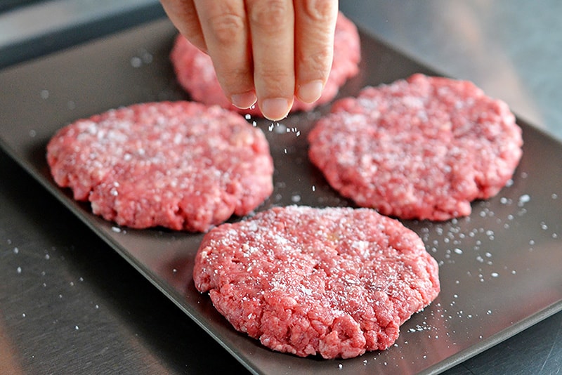 A hand is sprinkling kosher salt on four Nomster Burgers.