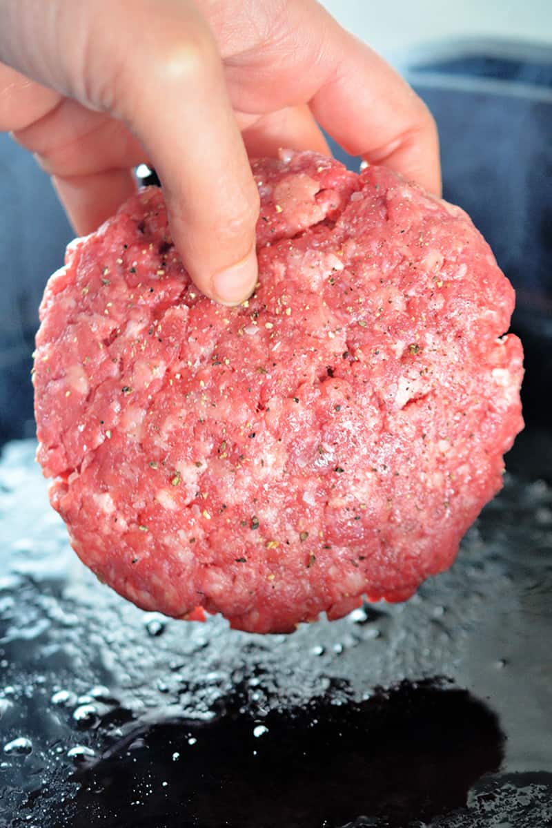 A hand is placing a Nomster Burger in a hot cast iron skillet.