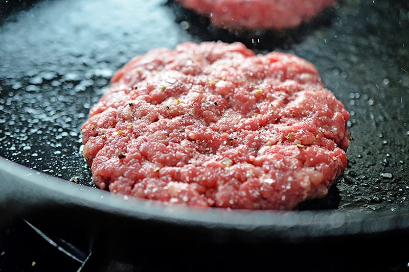 A closeup of Nomster burgers frying in a cast iron skillet with salt and pepper on the top.