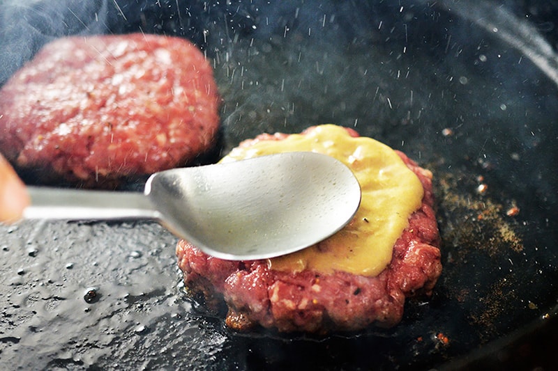 A spoon is spreading mustard on top of a Nomster burger frying in a cast iron skillet.