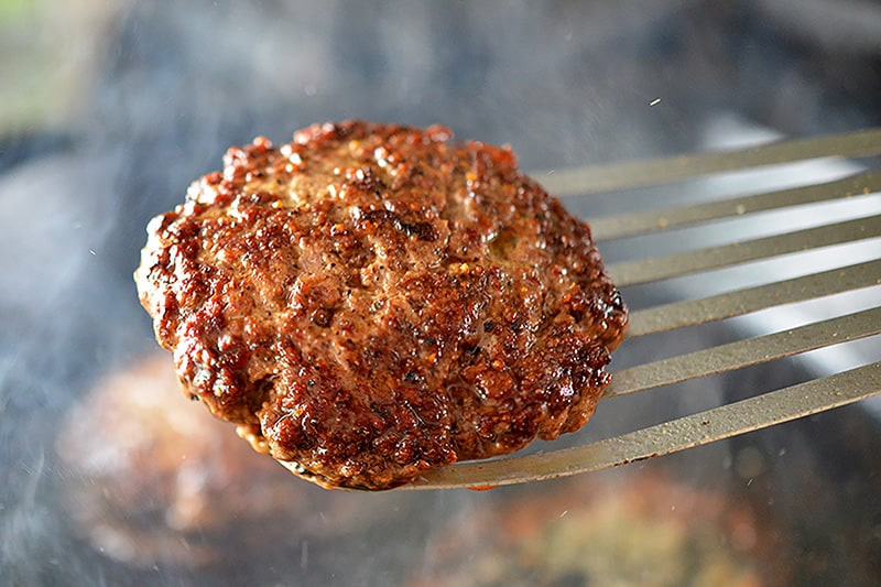 A cooked Nomster burger is being lifted out of a cast iron skillet with a metal fish spatula.