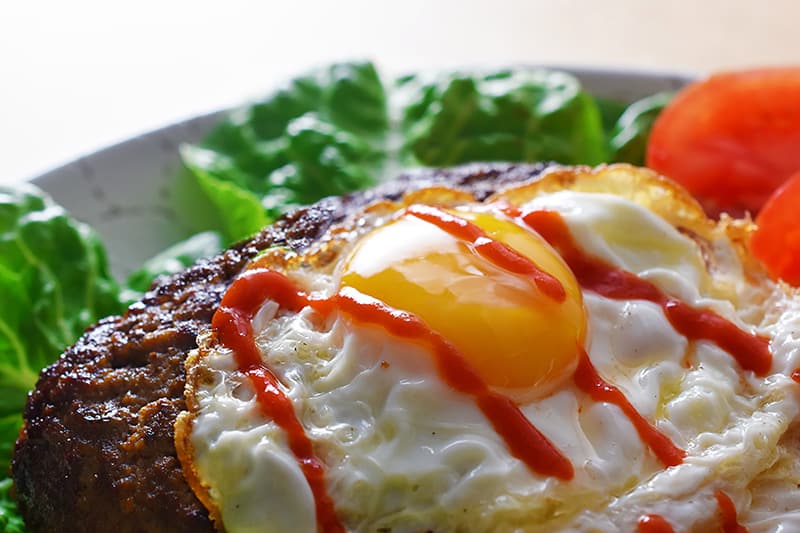 A side view of a Nomster burger on a bed of lettuce, topped with a crispy sunnyside egg, and a squiggle of sriracha.