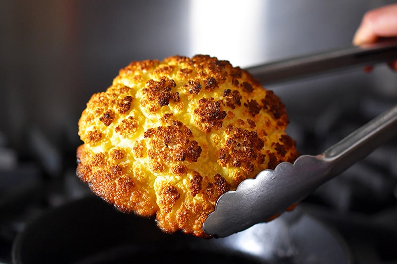 A pair of tongs is removing a Roasted Whole Cauliflower head from a cast iron skillet.