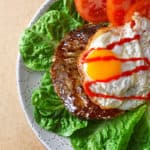 An overhead shot of a Nomster Burger on a bed of lettuce and topped with a crispy fried egg, sriracha, and sliced tomatoes.