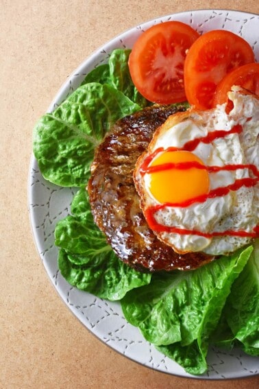 An overhead shot of a Nomster Burger on a bed of lettuce and topped with a crispy fried egg, sriracha, and sliced tomatoes.
