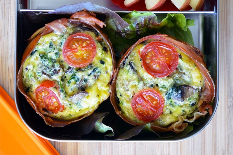 An overhead shot of two prosciutto wrapped frittata muffins in an open LunchBots bento box.