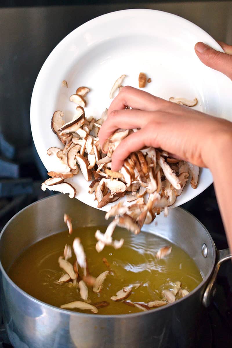 A person is adding a platter of sliced shiitake mushrooms to a saucepan filled with broth.