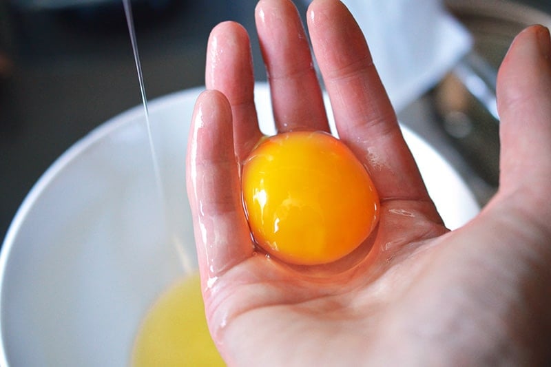 A hand holding a raw egg yolk on top of a white bowl filled with egg whites.