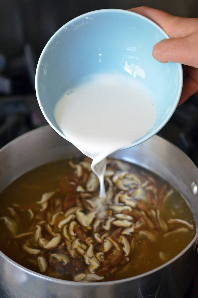 Pouring arrowroot slurry into a saucepan filed with simmering broth and sliced shiitake mushrooms