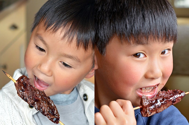 Two smiling Asian boys are eating paleo beef kabobs with Whole30 Cherry BBQ sauce
