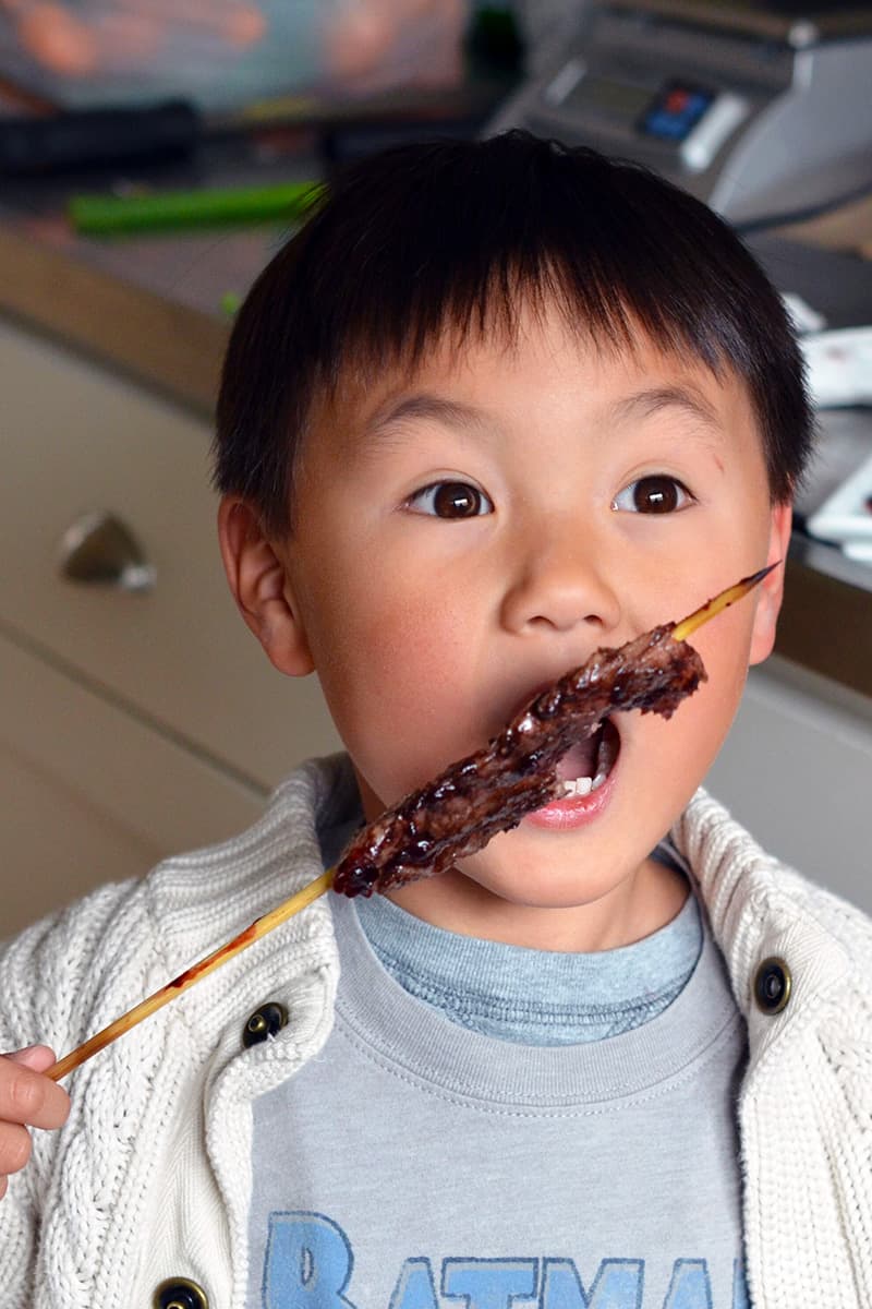 A little Asian boy is biting into a grilled beef kabob with Whole30 BBQ sauce