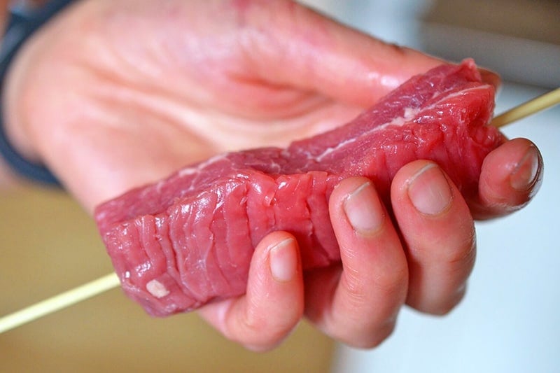 A hand is holding a piece of flank steak that has been skewered by a soaked bamboo skewer.