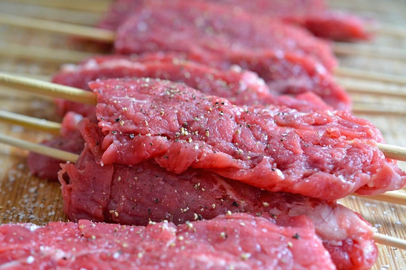 Flattened steak skewers are seasoned with salt and pepper .