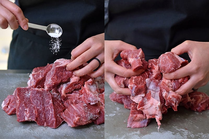 A shot of someone tossing cubed chuck roast with kosher salt on a gray cutting board.