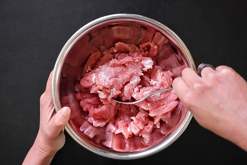 An overhead shot of two hands mixing sliced pork tenderloin strips with salt and avocado oil in a silver bowl.