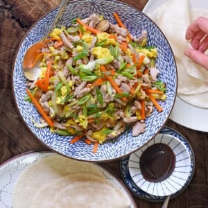 An overhead shot of a serving bowl filled with Paleo Moo Shu Pork, along with grain-free tortillas, and paleo hoisin sauce.