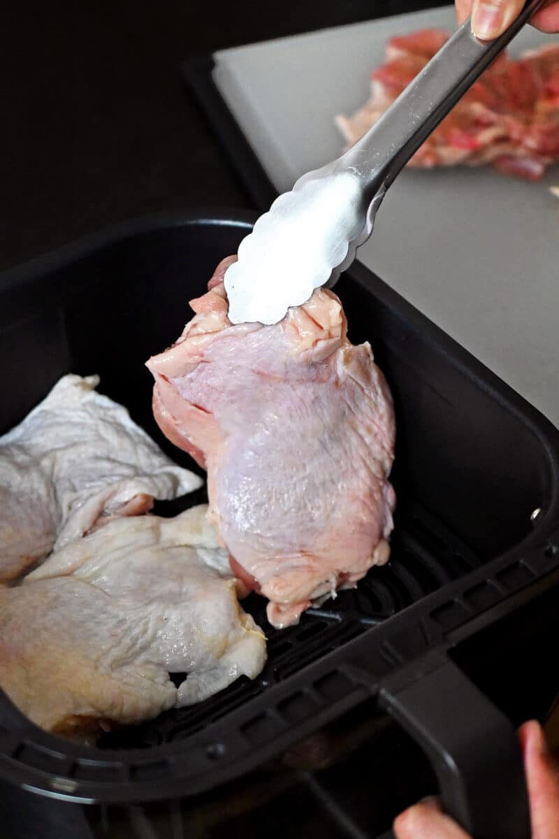 A pair of tongs is placing a raw chicken thigh into an air fryer basket.
