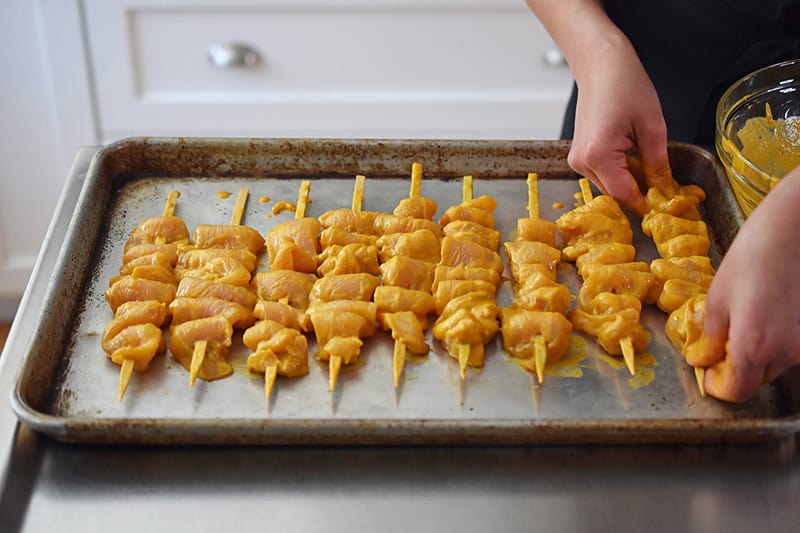 Placing the raw chicken satay skewers on a rimmed baking sheet before baking in the oven.
