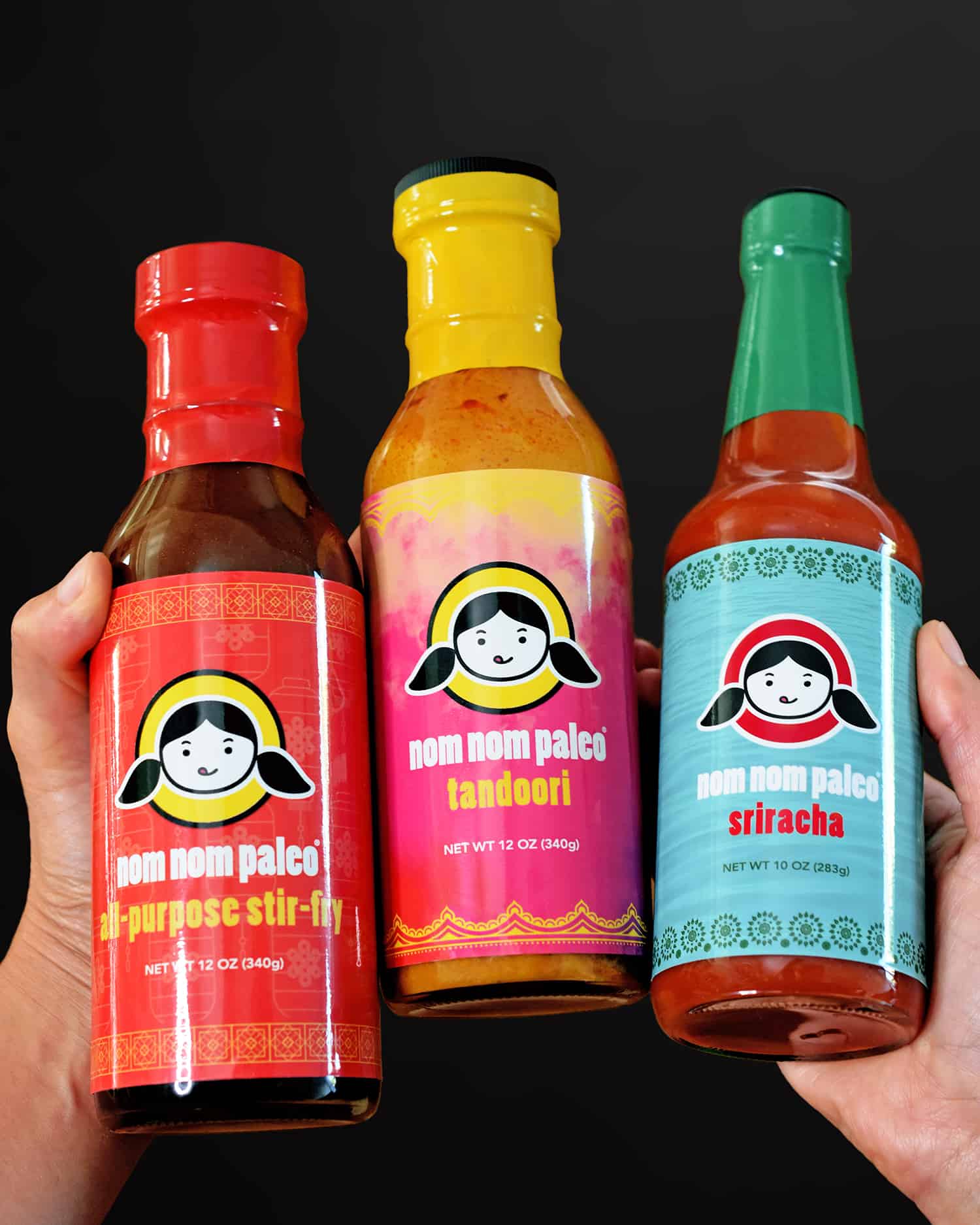Two hands are holding up three bottles of Nom Nom Paleo sauces: All-Purpose Stir-Fry, Tandoori, and Sriracha!