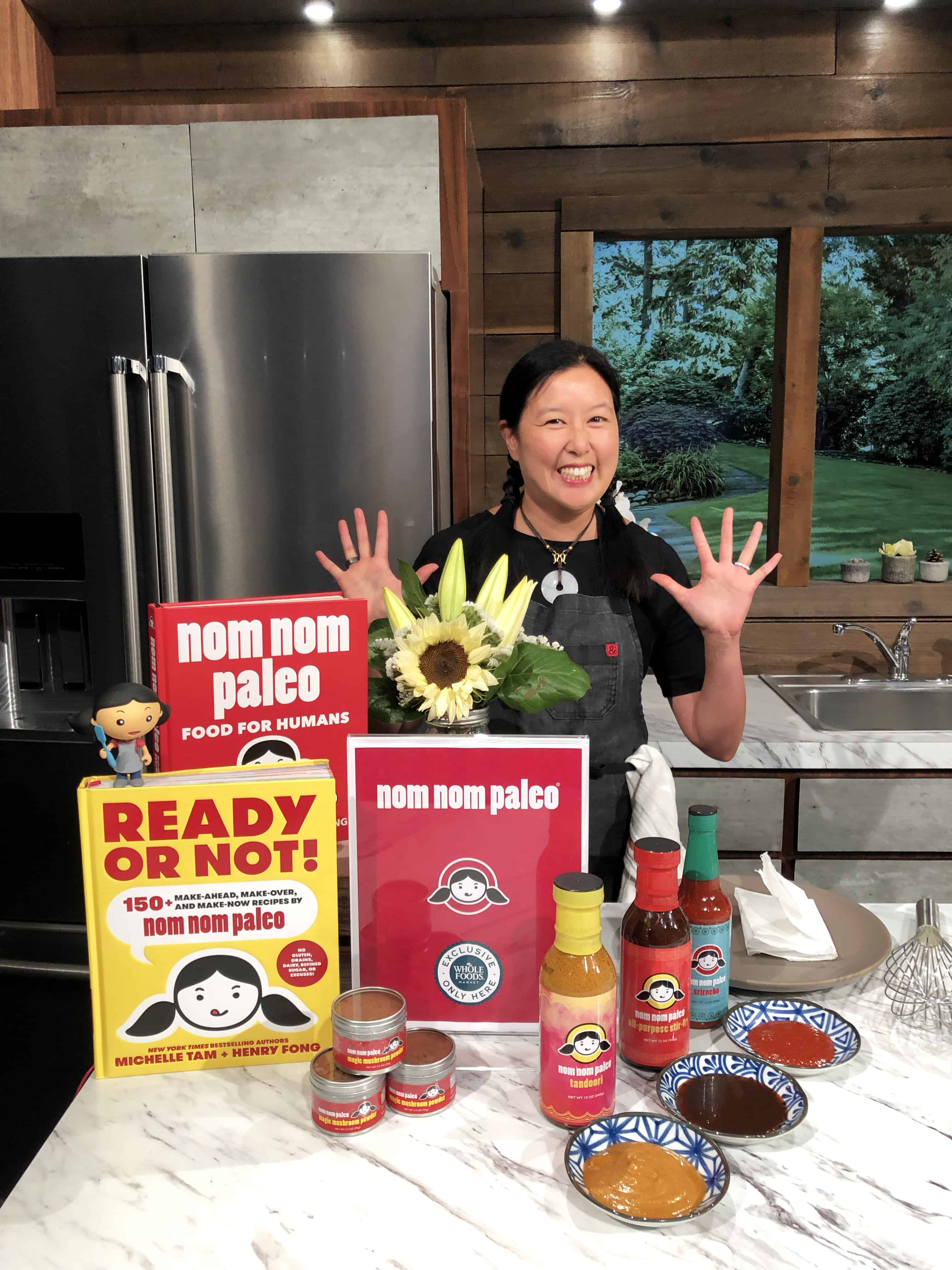 Michelle Tam standing in a kitchen behind a display of her Nom Nom Paleo cookbooks, sauces, and Magic Mushroom Powder.