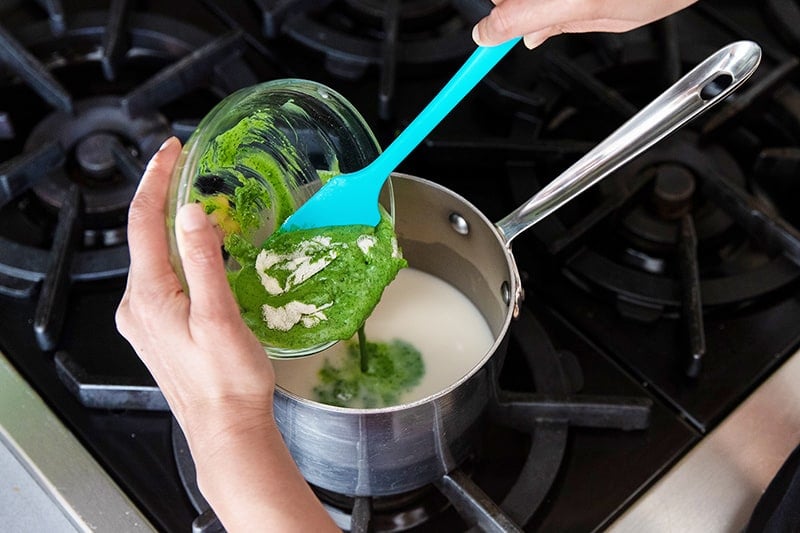 A blue silicone spatula spoons the matcha and gelatin mixture into the heated coconut milk.