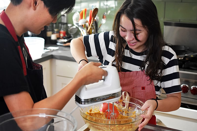 A teenage boy and a teenage girl are smiling as they are mixing a batch of paleo pumpkin muffins in a glass bowl.