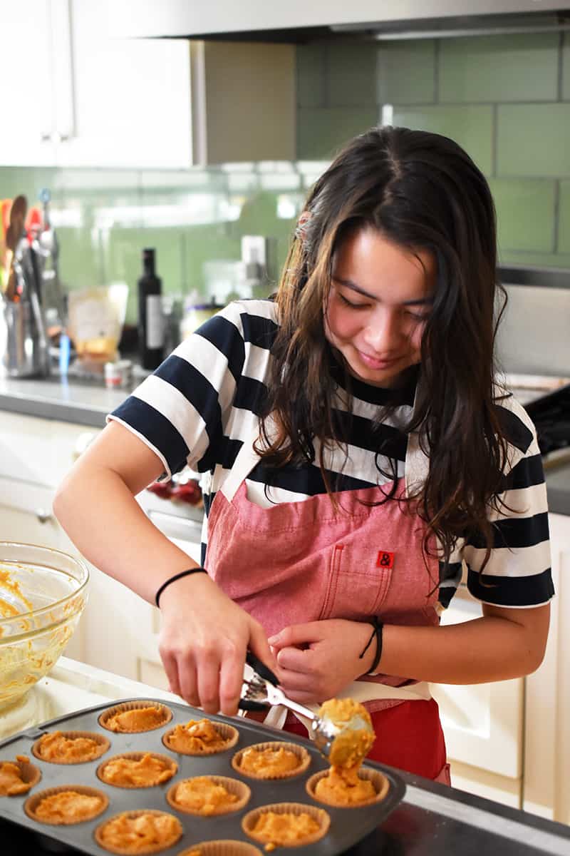 A teenage girl smiles as she scoops batter into a muffin tin.