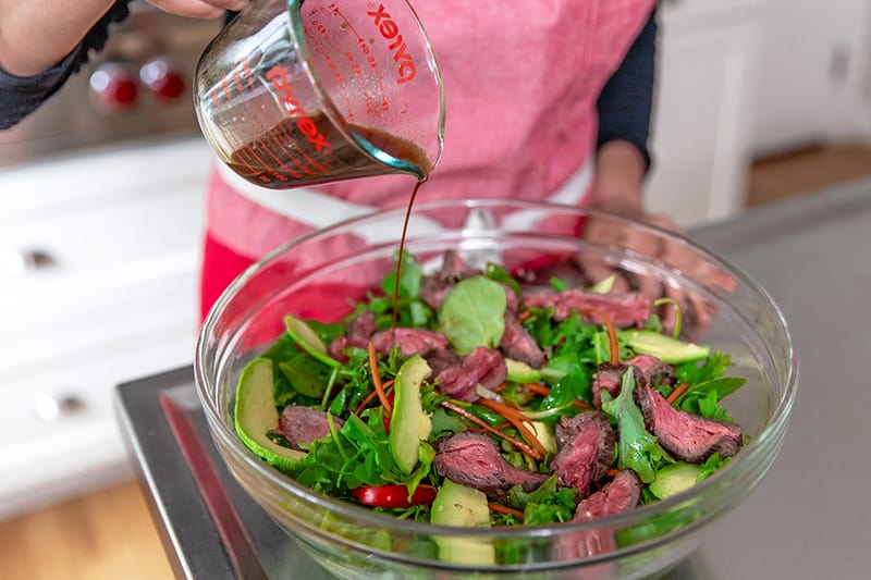 Pouring salad dressing on the Asian Steak salad that has sliced steak, avocados, and vegetables in a clear bowl.