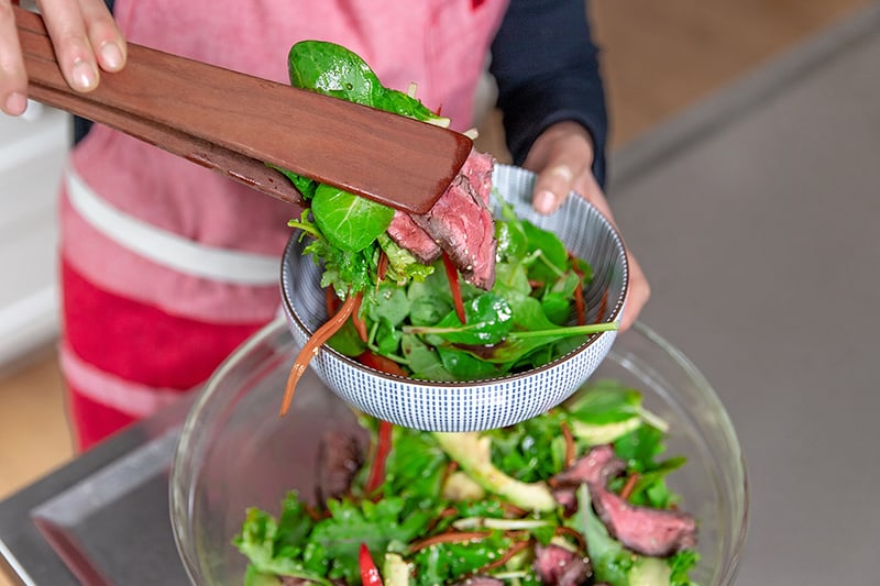 Using wooden tongs to place Asian Steak Salad into a bowl.