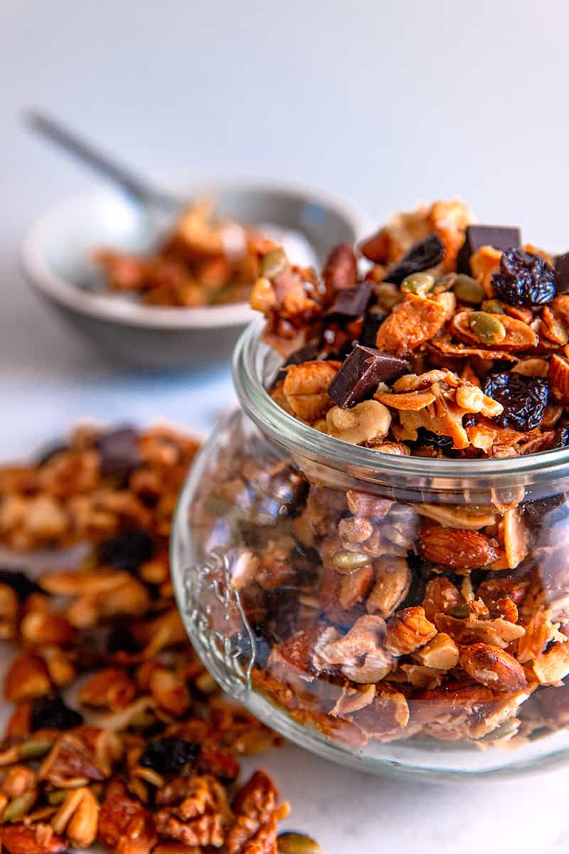 A closeup of a jar filled with Chocolate Cherry Paleo granola and a bowl in the background with granola on top of coconut yogurt.