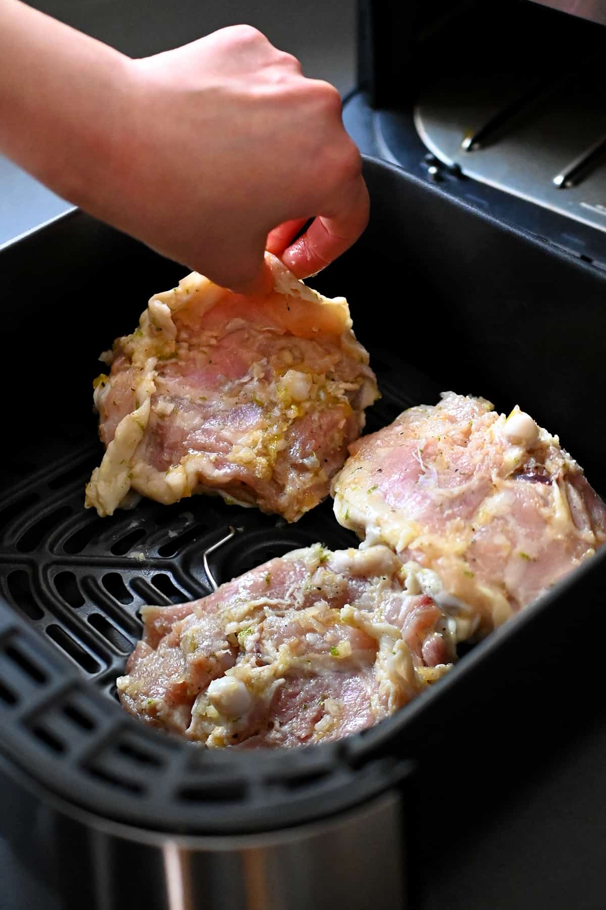 A hand is placing raw lemongrass chicken thighs in an open air fryer., skin side down.
