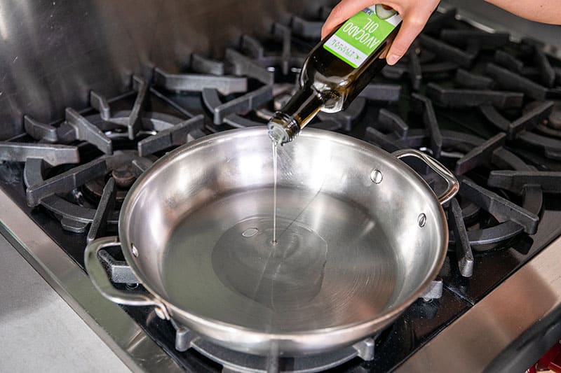 Pouring avocado oil from a bottle into a large stainless steel skillet.