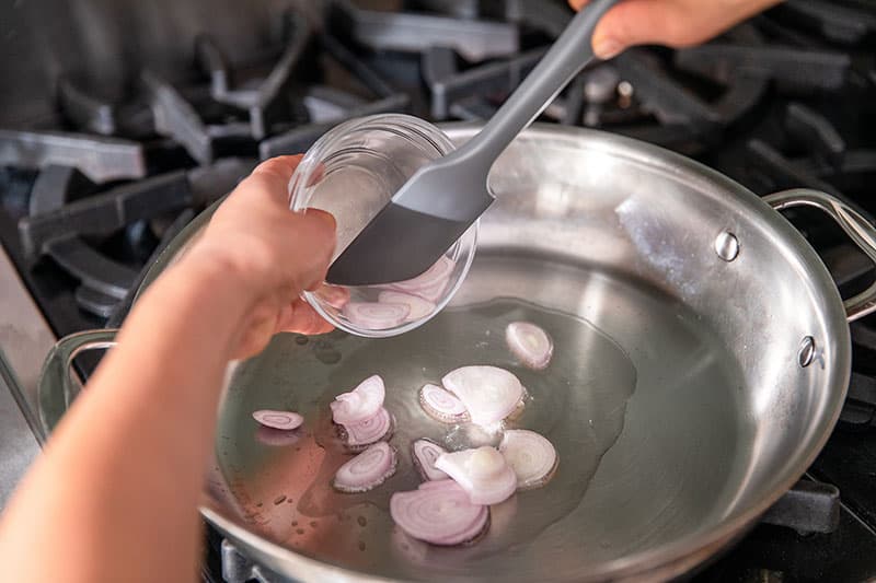 Adding thinly sliced shallots from a clear bowl to a hot stainless steel skillet.