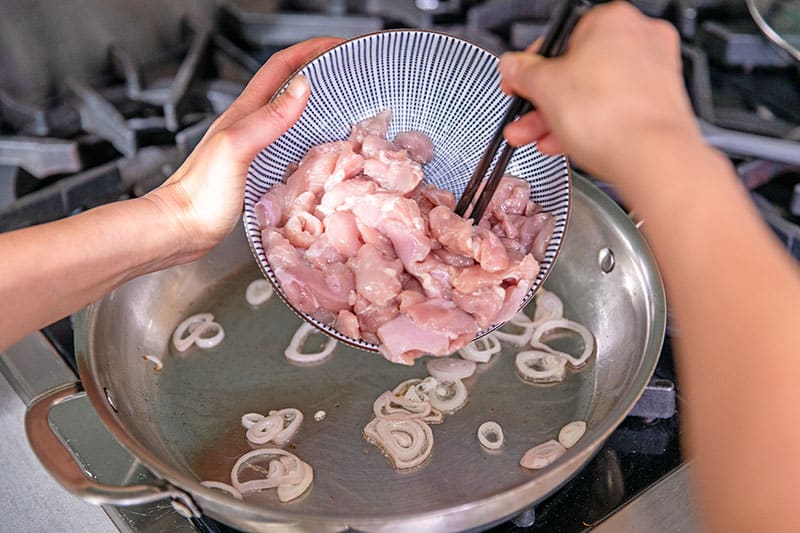 Adding sliced raw chicken thighs to a large stainless steel skillet with fried shallots in it.