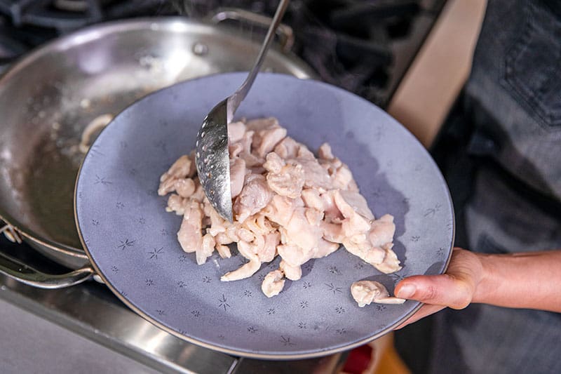 A slotted metal spoon is removing cooked stir-fried chicken out of a pan an onto a blue plate.