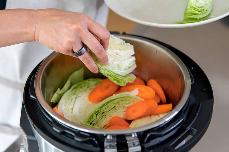 A hand is adding cabbage wedges and sliced carrots to an Instant Pot filled with Whole30 pork stew.