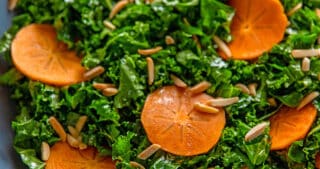 An overhead shot of two hands holding a large blue bowl filled with kale salad and persimmons and toasted almonds, a simple and delicious winter salad.