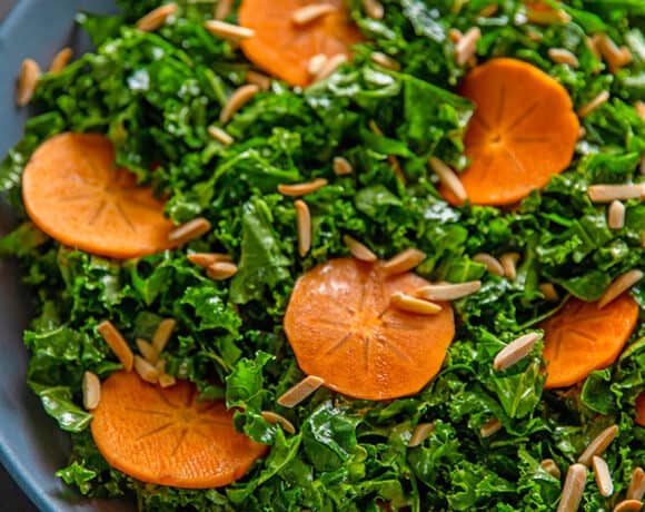 An overhead shot of two hands holding a large blue bowl filled with kale salad and persimmons and toasted almonds, a simple and delicious winter salad.