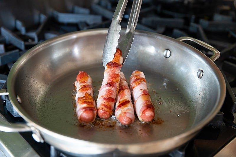 A pair of tongs are flipping golden brown bacon-wrapped Sonoran hot dogs in a large stainless steel skillet. 
