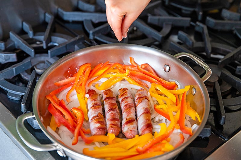 An overhead shot of a skillet with bacon-wrapped Mexican hot dogs surrounded by sliced onions and red and yellow bell peppers. A hand above is sprinkling salt on the vegetables.
