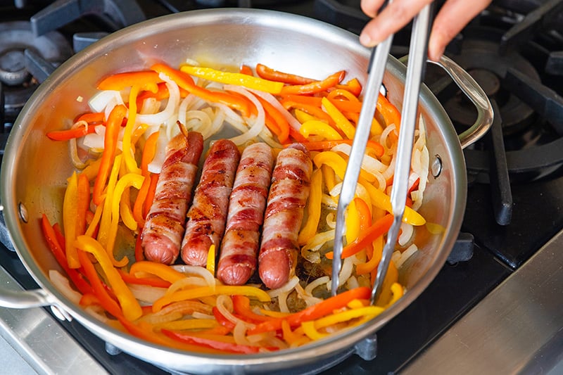 A shot of a skillet with browned bacon-wrapped Sonoran hot dogs with cooked sliced onions and peppers surrounding it. Tongs are moving the vegetables in the pan to cook them evenly.